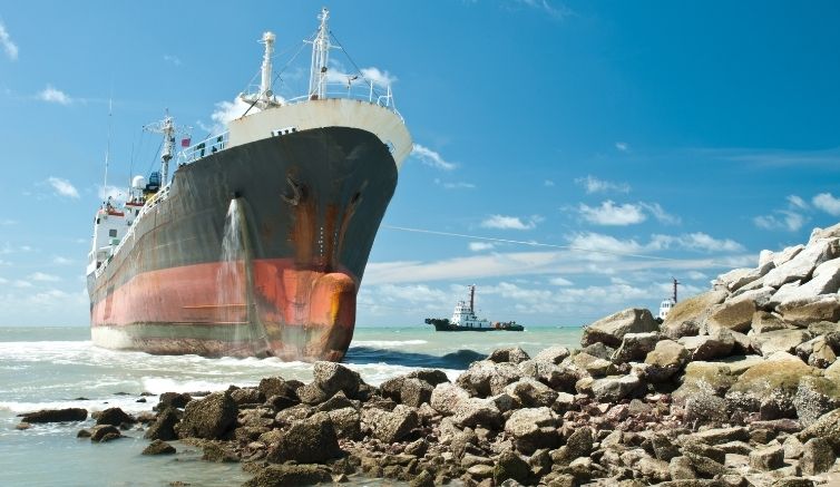 Large cargo ship run aground on a rocky coastline with waves crashing against the hull, illustrating the nautical origin of the idiom "on the rocks."